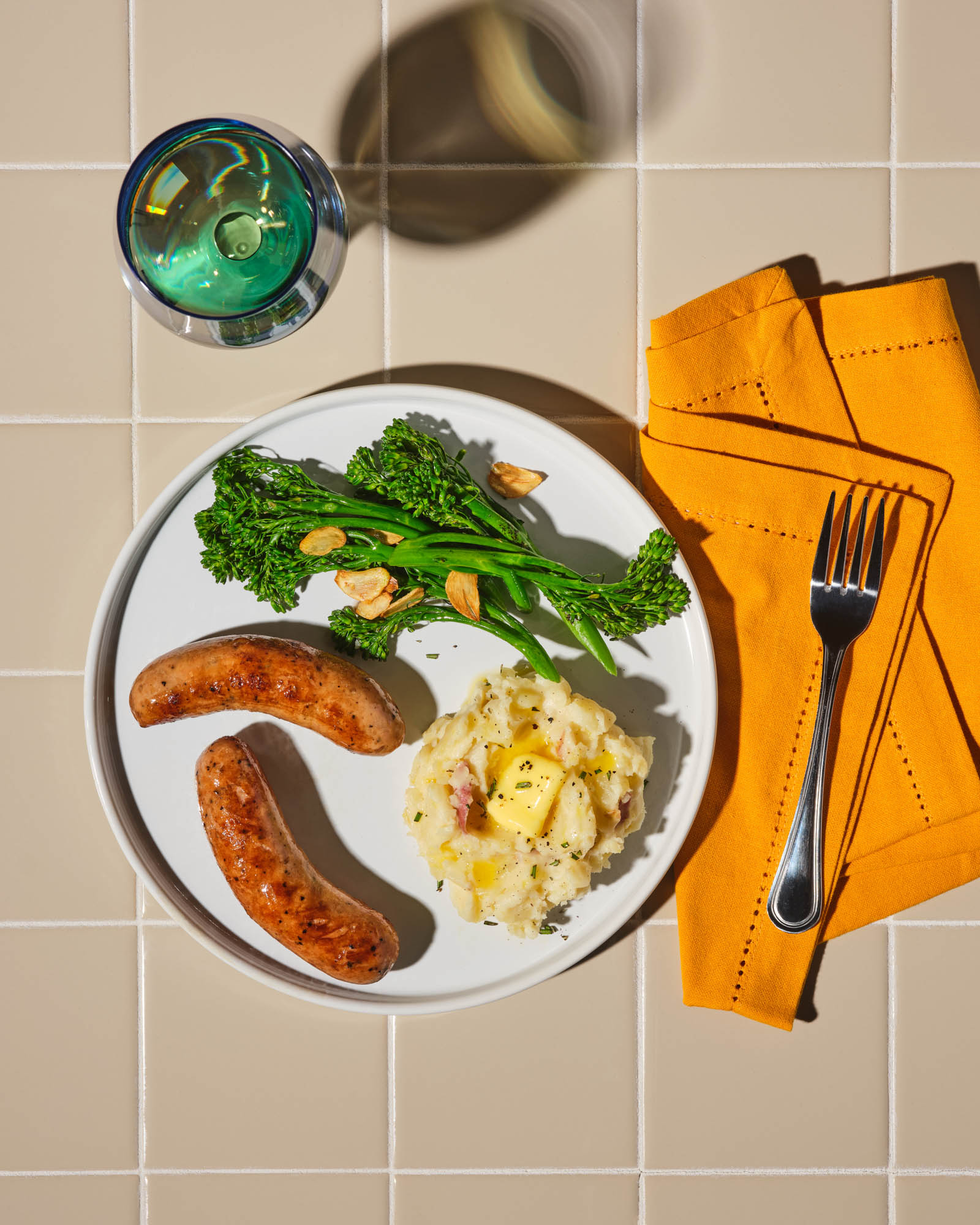 Grilled chicken sausage served with mashed potatoes, broccolini, and a drizzle of lemon butter sauce on a white plate.