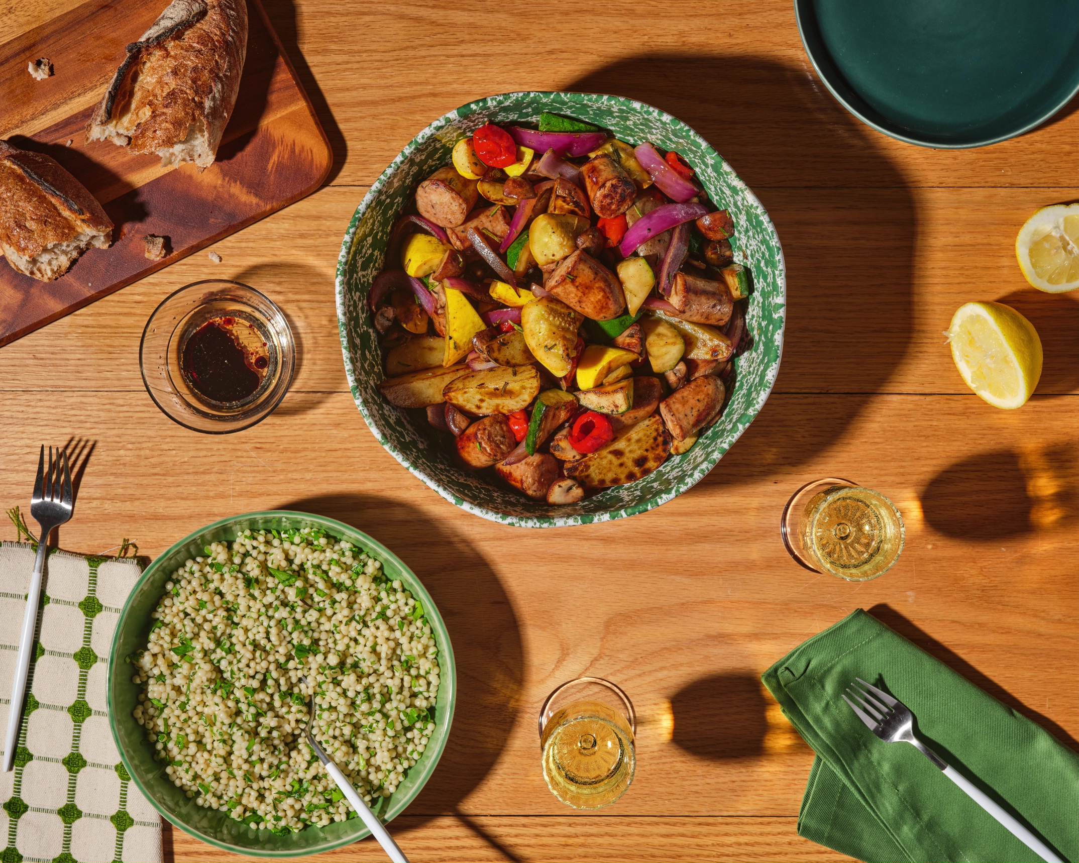 A dinner spread in which a bowl of The Sausage Project's Italian Herb Chicken Salad and roasted veggies is surrounded by quinoa, white wine, lemons, and rustic bread.