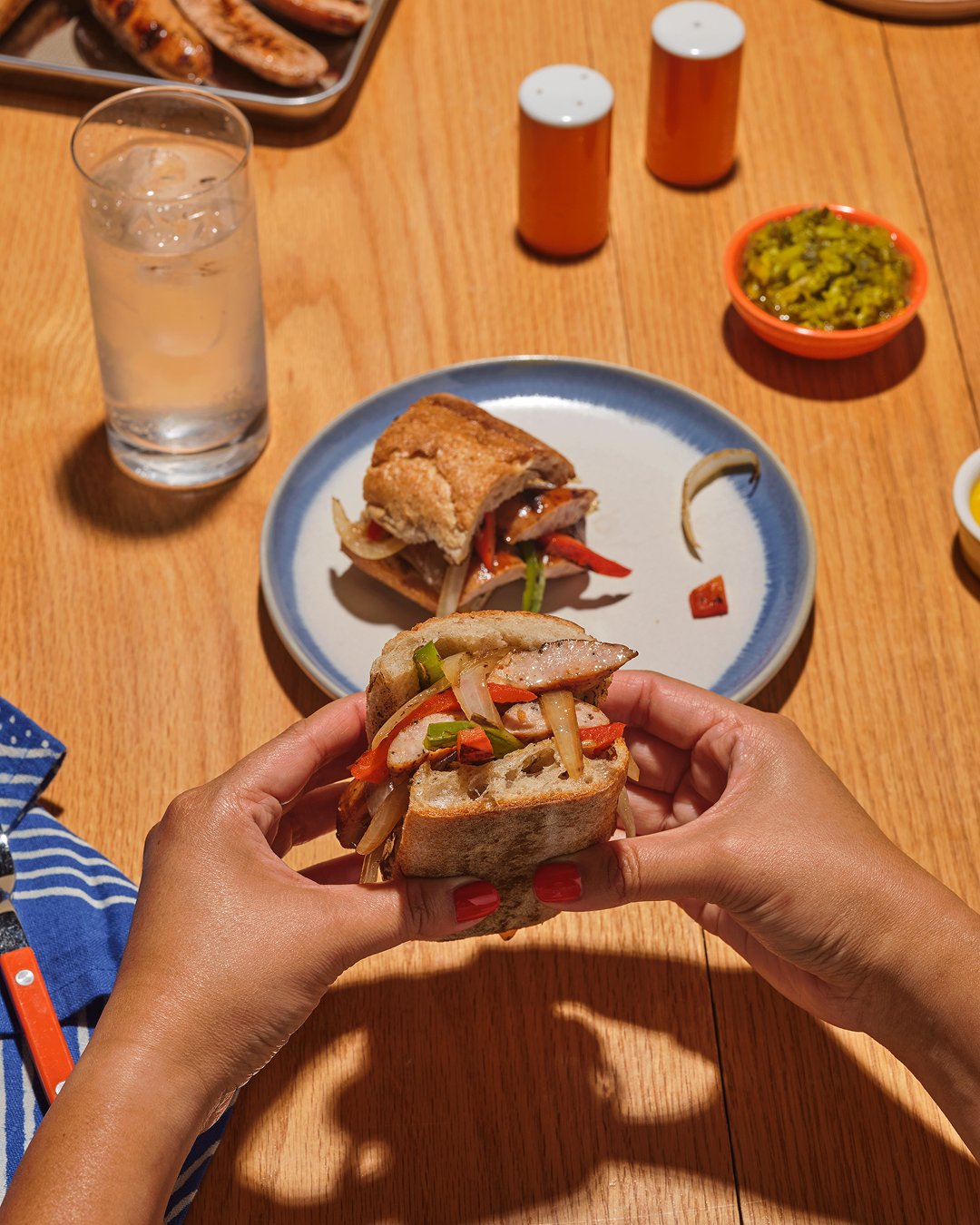 Hands holding a savory sausage, onion, and grilled pepper sandwich made with The Sausage Project's Italian Herb Chicken Sausage.