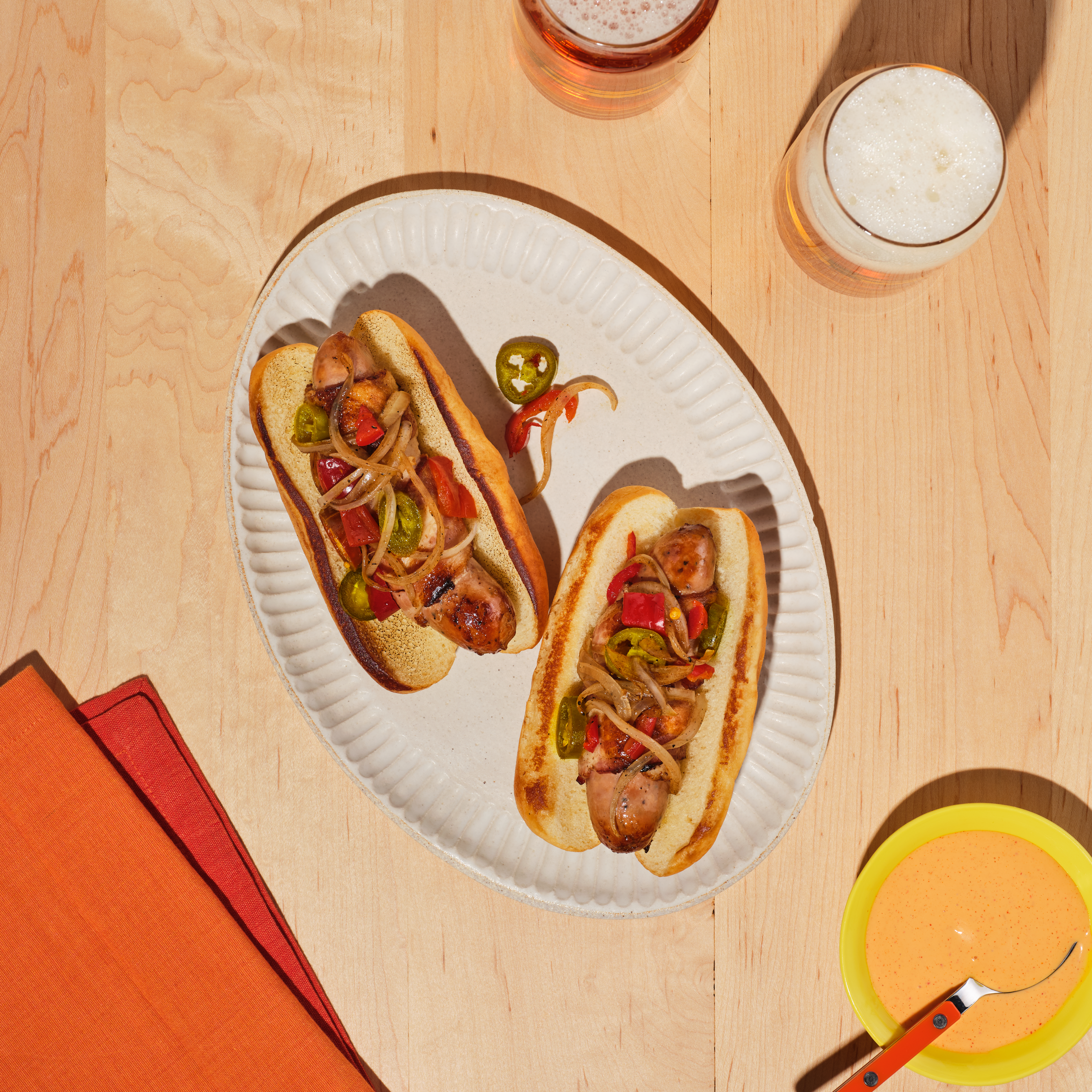 Bacon-wrapped Melty Cheddar Chicken Sausage topped with grilled peppers and onions on a toasted bun, served with a side of spicy gochujang aioli.