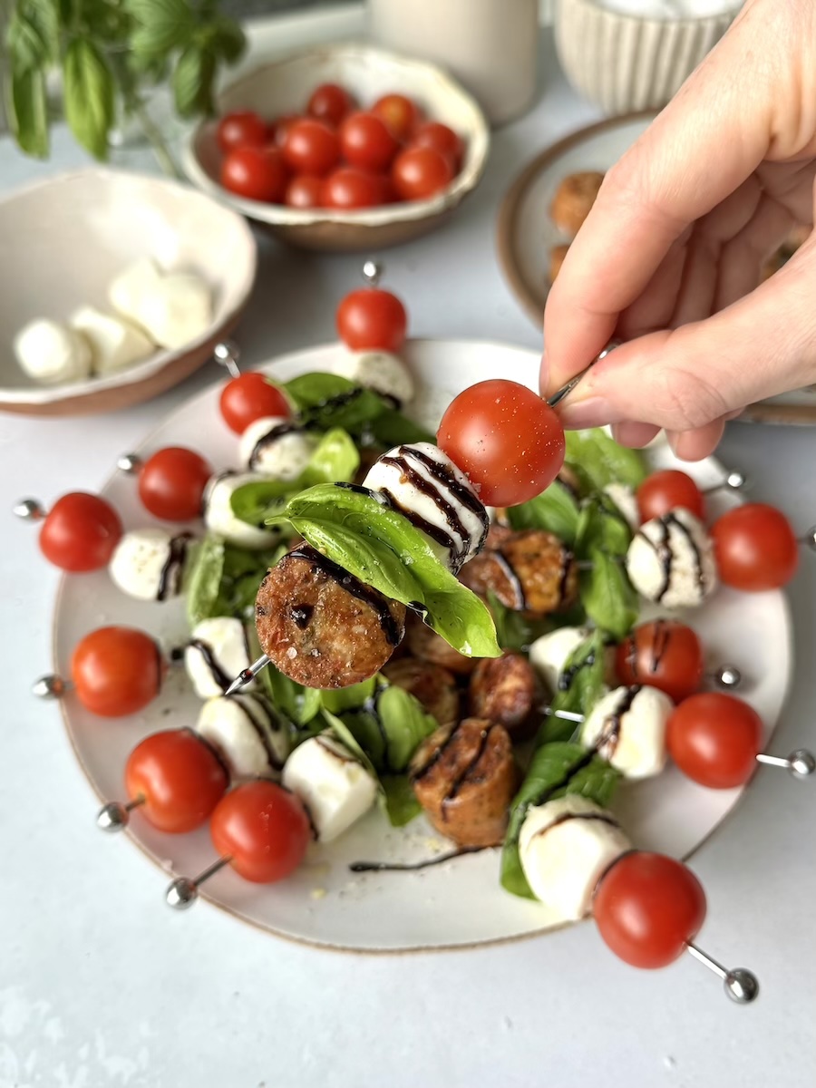 Hands grabbing a delicious caprese salad skewer featuring tomatoes, balsamic dressing, mozzarella, and Italian Herb Chicken Salad.