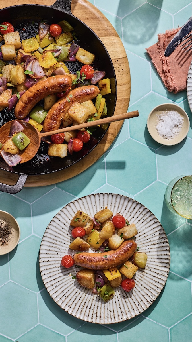 A skillet loaded with Melty Cheddar Chicken Sausage, potatoes, peppers, tomatoes, and onions and surrounded by a nicely made plate, drinks, and salt.