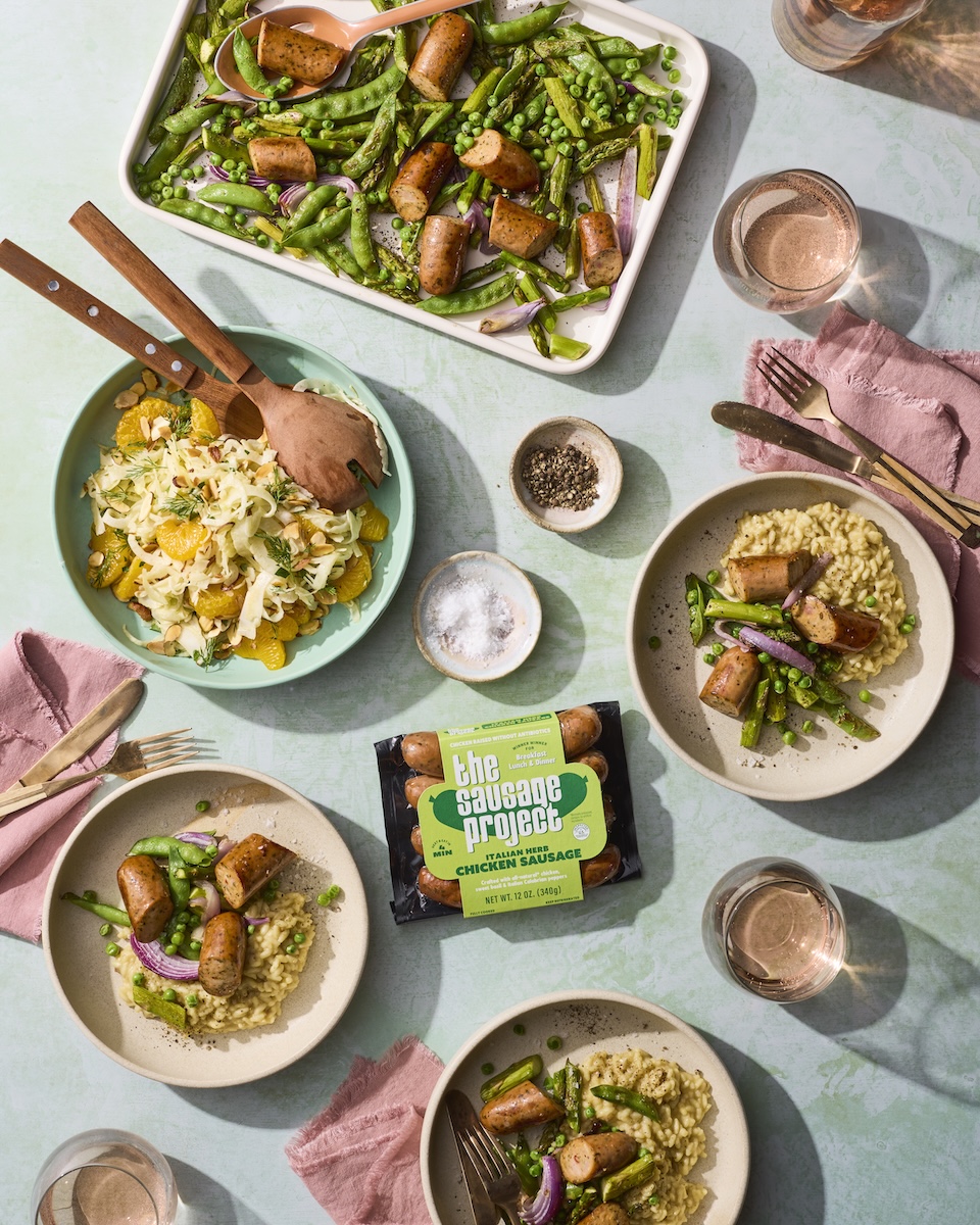A sheet pan full of sausage and vibrant vegetables on an elegant table setting beside a package of Italian Herb Chicken Sausage.